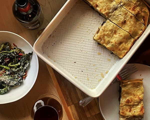 An overview shot of a half empty pan of lasagna, a slice sitting on a white plate beside it, with a bowl of greens off to the side, along with a bottle of red wine a glass of red wine.